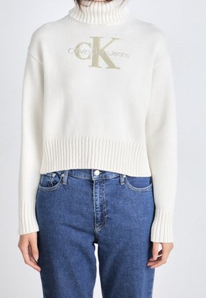 Kvinde iført en hvid rullekravetrøje med Calvin Klein Jeans-logo og blå jeans med høj talje.