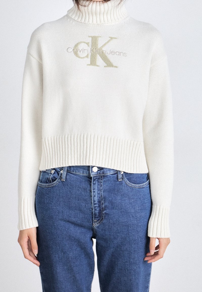 Femme portant un pull à col roulé blanc avec le logo Calvin Klein Jeans et un jean bleu taille haute.