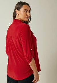 Rote Bluse mit langen Ärmeln, Kragen und hochrollbaren Ärmeln; glatte Textur; lässiger Schnitt; verfügt über eine einzelne Brusttasche auf der linken Seite.