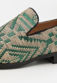 Chaussures à enfiler présentant un motif tissé beige et vert, avec un matériau texturé et une bordure noire. Talon bas noir, durable.