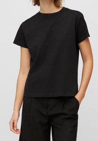 Persona vistiendo una camiseta de manga corta negra lisa y pantalones negros, de pie con una mano en el bolsillo frente a un fondo liso.