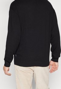 Zwart sweater met lange mouwen, geribbelde manchetten en zoom. Gemaakt van een zachte stof, met een subtiele labeldetail aan de onderkant links.