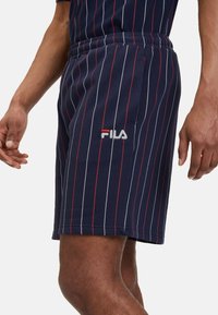 Pantaloni corti blu navy con strisce verticali rosse e bianche. Vita elastica con un logo Fila visibile in bianco sul lato. Tessuto leggero.