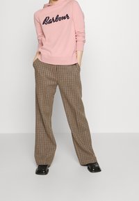 Różowa bluza z kapturem z granatowym logo „Barbour”, w zestawie z szerokimi, beżowo-brązowymi spodniami w kratę, wyposażonymi w boczne kieszenie i błyszczące czarne buty.