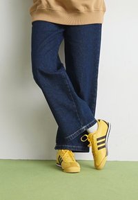 Geltoni sportiniai bateliai su juodomis juostelėmis, dėvimi su tamsiai mėlynomis plačiomis džinsomis. Audinys atrodo tvirtas, su nedideliu tekstūros jausmu ir nuplyšusiu kraštu.