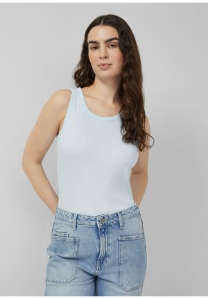 Femme aux longs cheveux bouclés portant un haut sans manches bleu clair et un jean en denim clair taille haute, posant devant un fond gris uni.