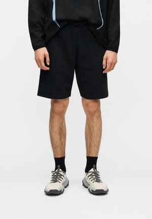 Άνδρας με μαύρο casual σορτς, μαύρες κάλτσες, μπεζ και γκρι αθλητικά παπούτσια, στέκεται μπροστά σε απλό φωτεινό φόντο.