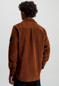 Camicia di velluto a coste marrone con colletto strutturato, polsini con bottone e spacco posteriore. Presenta una toppa nera sulla manica sinistra.