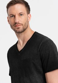 Černé tričko s výstřihem do V, které má jemnou texturu, krátké rukávy a kapsu na levé straně hrudi. Tkanina vypadá lehce a neformálně.