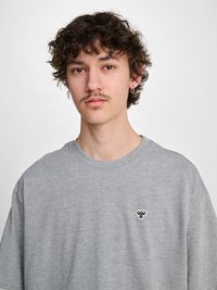 Jeune homme aux cheveux bouclés, moustache, portant un t-shirt gris avec un petit logo d'abeille sur la poitrine gauche, fond neutre.