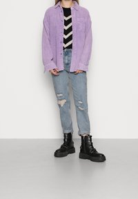 Chemise en velours côtelé violet clair, pull noir et blanc à motifs zigzag, jeans bleus clairs usés et bottes de combat noires épaisses avec des accents en métal.