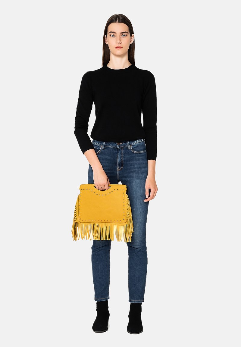 Femme jeune portant un top noir à manches longues, un jean bleu, des bottes noires, tenant un sac à franges jaune avec des rivets en métal, sur fond blanc.