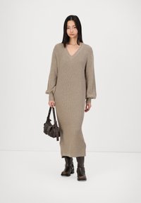 Stickad beige midiklänning med V-ringning, ballongsärmar och ribbad textur, tillsammans med en svart axelväska och bruna ankelstövlar.