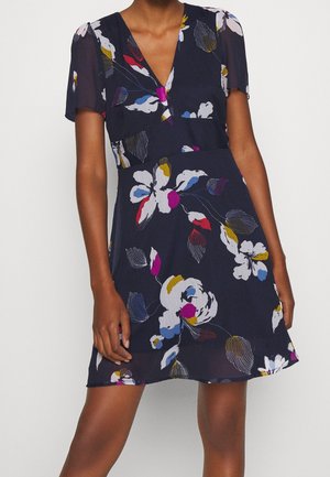 Femme portant une robe bleu marine à encolure en V avec des manches courtes, arborant un imprimé floral coloré en blanc, rose, rouge, bleu et jaune.