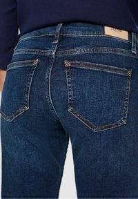 Jean en denim bleu foncé avec une texture lisse, présentant un design classique à cinq poches et des coutures orange contrastantes sur les coutures.