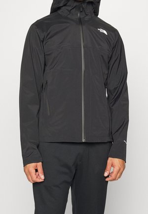 Mężczyzna noszący czarną kurtkę na zamek z kapturem i bocznymi kieszeniami na zamek, z logo The North Face na klatce piersiowej.