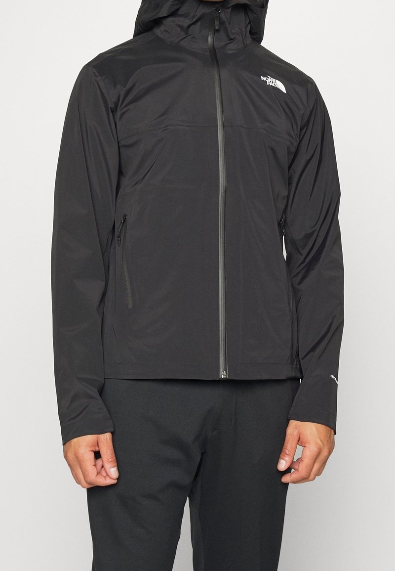 Homme portant une veste noire zippée avec capuche et poches latérales zippées, avec le logo The North Face sur la poitrine.