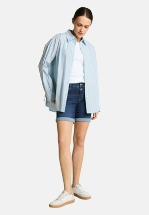 Frau trägt hellblau gestreiftes Hemd mit Knöpfen, weißes Top, hochgekrempelte Jeansshorts und weiße Sneaker, steht vor einem einfarbigen Hintergrund.