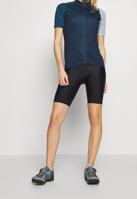Marineblaues Fahrradtrikot mit kurzen, gemusterten Ärmeln, kombiniert mit schwarzen Fahrradhosen. Verfügt über einen Frontreißverschluss und strukturierte Details an den Ärmeln.