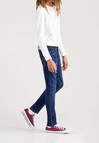 Mörkblå denimjeans med slim fit, ihop med en vit långärmad skjorta och vinröda låga sneakers. Har synliga sömmar och fickor.