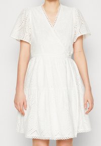 Robe blanche en coton avec un design portefeuille, des manches courtes bouffantes, un motif en tissu ajouré et un ourlet festonné.