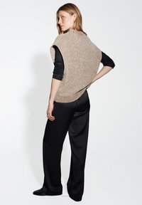 Maglione senza maniche beige in maglia con una finitura testurizzata, indossato sopra una maglietta nera a manica lunga, abbinato a pantaloni neri a gamba larga.