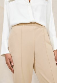 Lichtbeige op maat gemaakte broek met een gladde textuur, twee voorplooien en een hoge tailleband, gecombineerd met een wit overhemd met knopen.