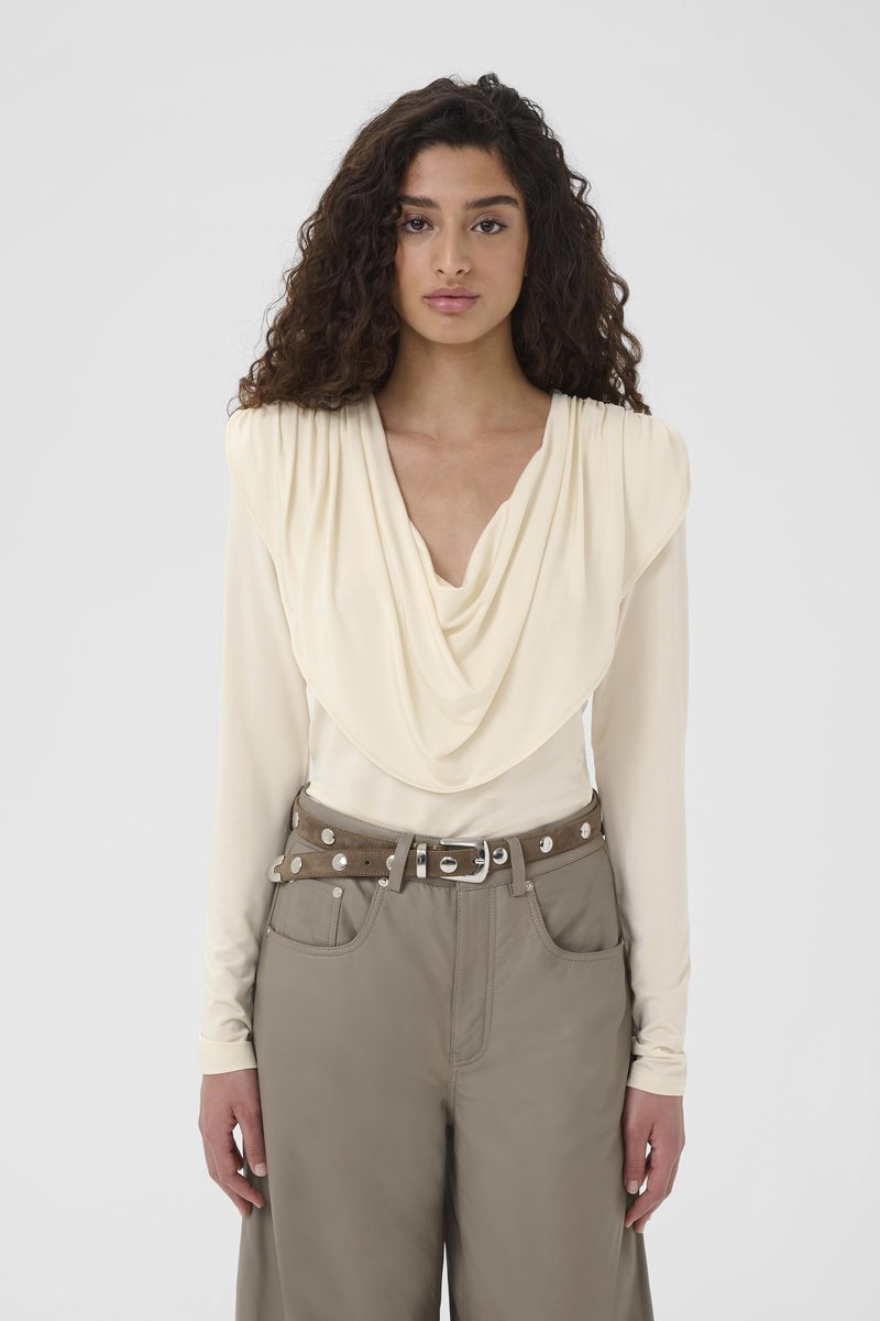 Noor naine lokkis juustega, kes kannab kreemikaid volditud kaelusega pluusi ja halla toonides laiade säärtega pükse koos naastustega pruuni vööl, seisab valge tausta ees.