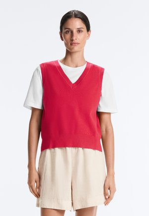 Femeie purtând un vestă tricotată fără mâneci, roșie cu decolteu în V, peste o cămașă albă cu mâneci scurte și pantaloni scurți bej, stând în fața unui fundal alb.