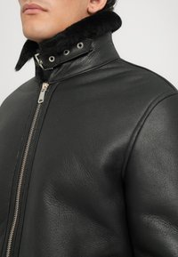 Bărbat îmbrăcat într-o jachetă din piele neagră cu guler pufoș, având un fermoar argintiu, arătând un prim-plan al zonei superioare a pieptului și gâtului.