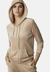 Hoodie zippé en velours beige avec capuche, poches avant et poignets côtelés. Texture lisse avec un aspect doux et pelucheux. Détail de fermeture éclair dorée.