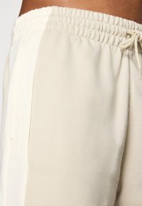 Kremfargede joggebukser med elastisk midje, snor og sidelommer laget av myk, lettvektig stoff.