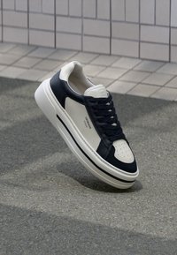 Sneaker mit einem oberteil aus marineblauem Leder und cremefarbenen Akzenten, ausgestattet mit einer durchlöcherten Zehenpartie, flachen Schnürsenkeln und einer strukturierten Gummisohle.