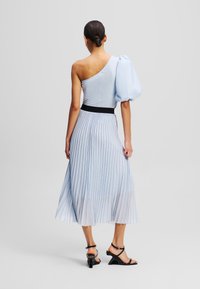 Robe légère bleu clair à une épaule avec une manche bouffante, jupe plissée, ceinture noire et texture lisse. Des sandales à talons complètent le look.