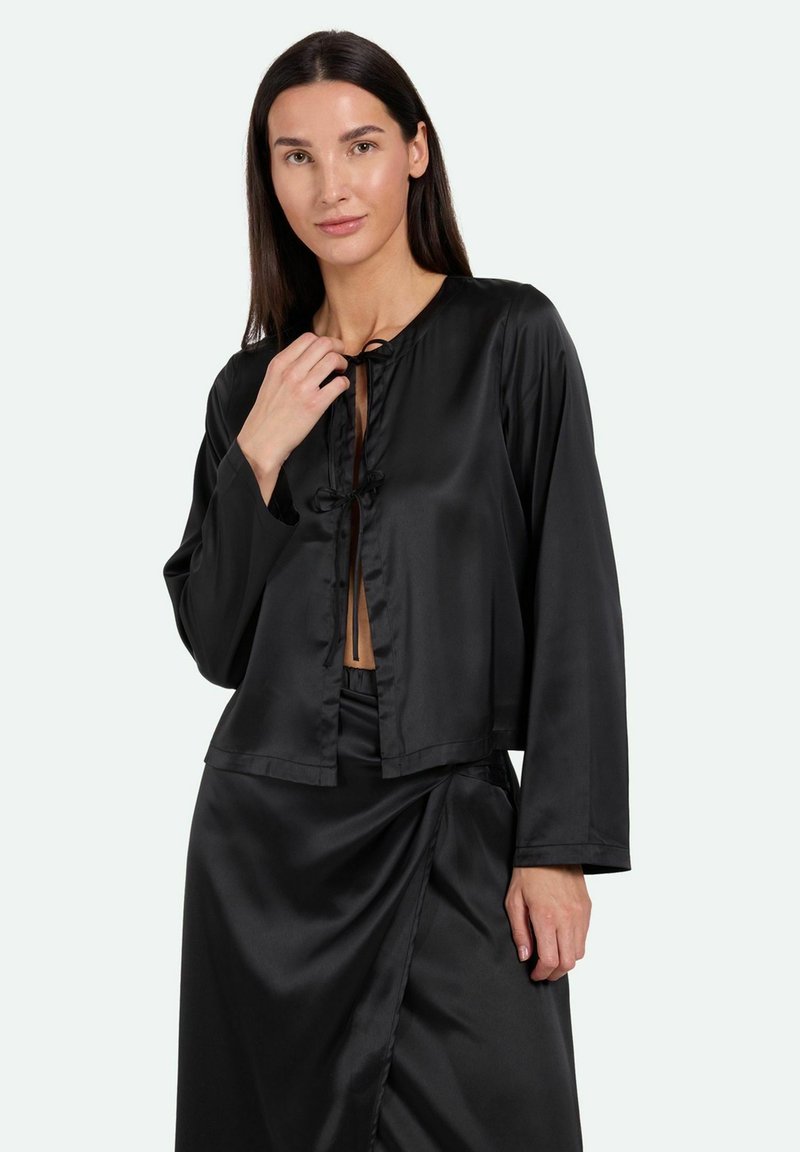 Schwarze Satin-Crop-Jacke mit langen Ärmeln, rundem Hals und gebundenem Frontverschluss. Glatte Oberfläche, schlichtes Design, kombiniert mit passenden Hosen.