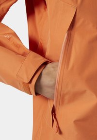 Chaqueta impermeable naranja con un bolsillo lateral con cremallera, textura suave y puños ajustables con cierre de Velcro. Diseño ligero y transpirable.