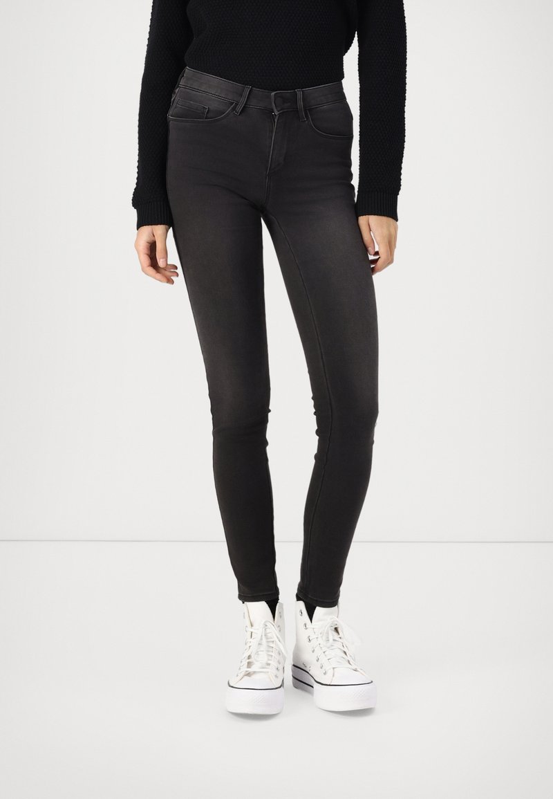 Jeans negros ajustados con una textura suave, cintura alta, cinco bolsillos y trabillas para el cinturón, combinados con zapatillas blancas de caña alta.