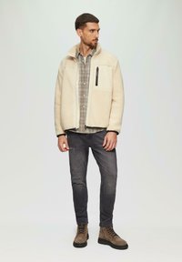 Veste polaire crème avec une poche zippée, superposée sur une chemise à carreaux grise. Associée à un jean foncé et des bottes à lacets marron.