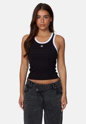 Kobieta o długich brązowych włosach, ubrana w czarny top bez rękawów z białym obszyciem oraz czarne jeansy z wysokim stanem, stojąca na tle jednolitego tła.