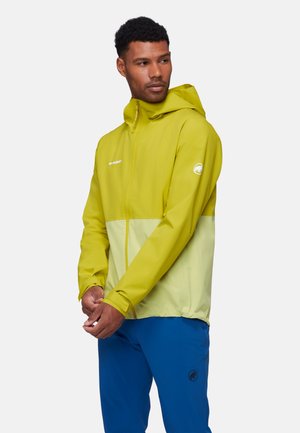 Man draagt een geel tweekleurig jack met capuchon en blauwe broek, kijkt opzij met handen voor zich gevouwen.