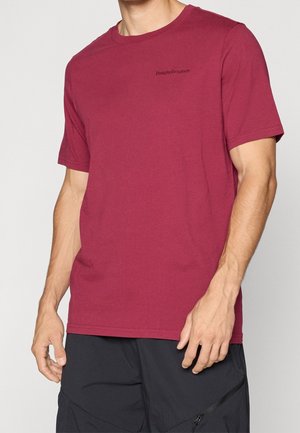 Mann trägt ein schlichtes weinrotes T-Shirt mit kurzen Ärmeln und kleinem schwarzem "Peak Performance"-Schriftzug auf der Brust und schwarze Hose.