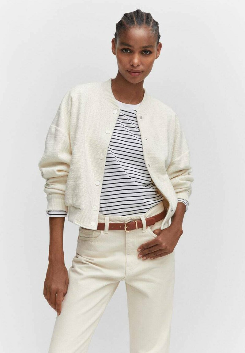 Frau in einem weißen strukturierten, knopfartigen Cardigan über einem schwarz-weißen gestreiften Hemd, beigen Hosen und einem braunen Gürtel vor einem einfarbigen Hintergrund.