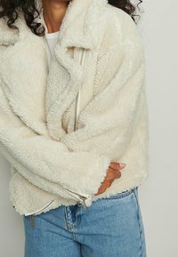Chaqueta esponjosa color crema con una textura suave, que presenta un cuello, cierre con cremallera y puños con cremallera. Usada sobre unos jeans de color azul claro.