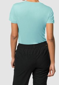 Bakifrån vy av en person i en ljusblå kortärmad skjorta och svarta elasticerade midjebandbyxor mot en enhetlig bakgrund.