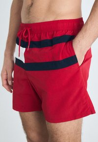 Rote Badehose mit Kordelzugbund, ausgestattet mit horizontalen Streifen in Marineblau und Weiß. Hergestellt aus leichtem, glattem Material mit Seitentaschen.
