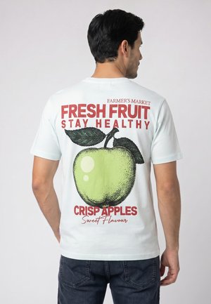 Mann trägt weißes T-Shirt mit großem grünem Apfel und dem Text "Fresh Fruit Stay Healthy Crisp Apples Sweet Flavour" auf dem Rücken.