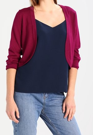 Femme portant un haut sans manches bleu marine à décolleté profond, un cardigan court violet et un jean bleu clair sur un fond uni.