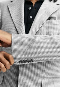 Gray and white houndstooth blazer with a structured fit, featuring a black button-up shirt underneath, and four black buttons on the cuff.