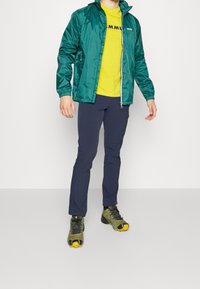 Veste en nylon vert avec une fermeture éclair, t-shirt jaune avec un texte noir, pantalon marine et chaussures de randonnée vertes avec des semelles jaunes.