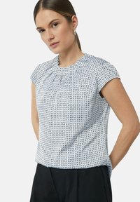 Kurzarmoberteil mit einem hellblauen Muster, das geometrische Formen zeigt. Strukturierter Stoff mit Raffung am Hals. Kombiniert mit schwarzen Hosen.